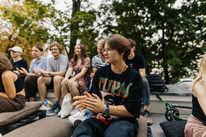 Jauna mergina Gabrielė, apsirengusi laisvu stiliumi, sėdi lauke, apsupta draugų.