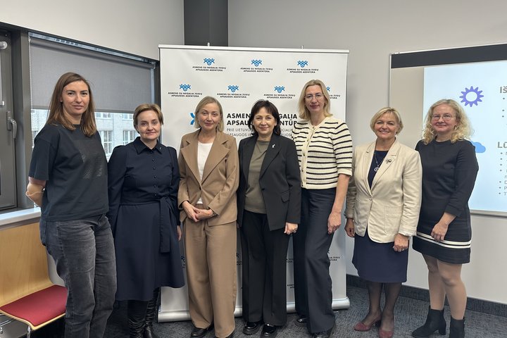 Prie foto sienelės su Agentūros logotipas stovi septynios moterys, Agentūros direktorės pavaduotoja Jelena Ivančenko, socialinės apsaugos ir darbo viceministrė Rita Grigalienė, Agentūros vadovė Eglė Čaplikienė, socialinės apsaugos ir darbo ministrė Jūratė