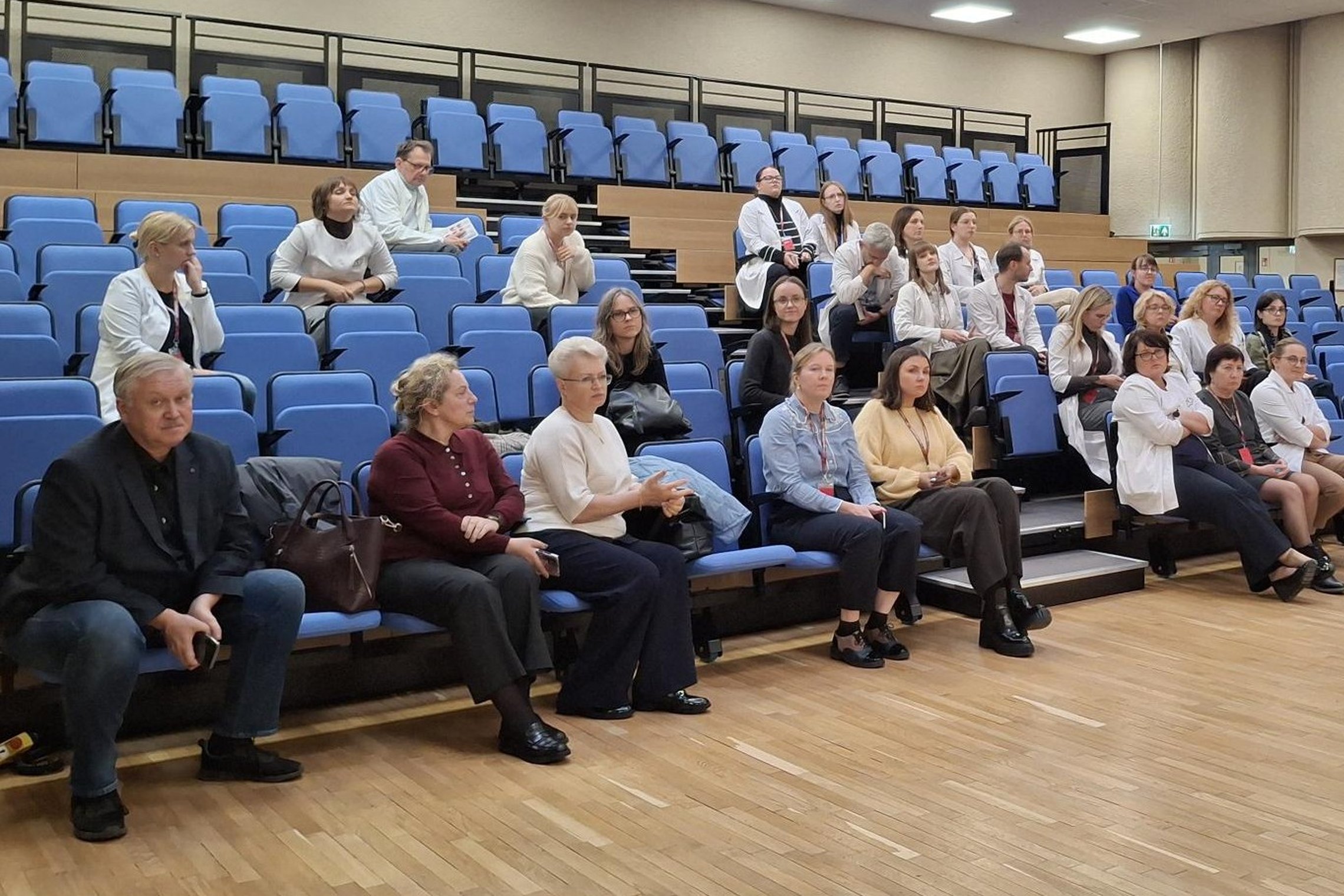 Salėje keliomis į viršų kylančiomis eilėmis sėdi įvairūs pranešimo klausytojai, gydytojai ir kiti gydymo įstaigų specialistai, dauguma jų dėvi baltus chalatus. 