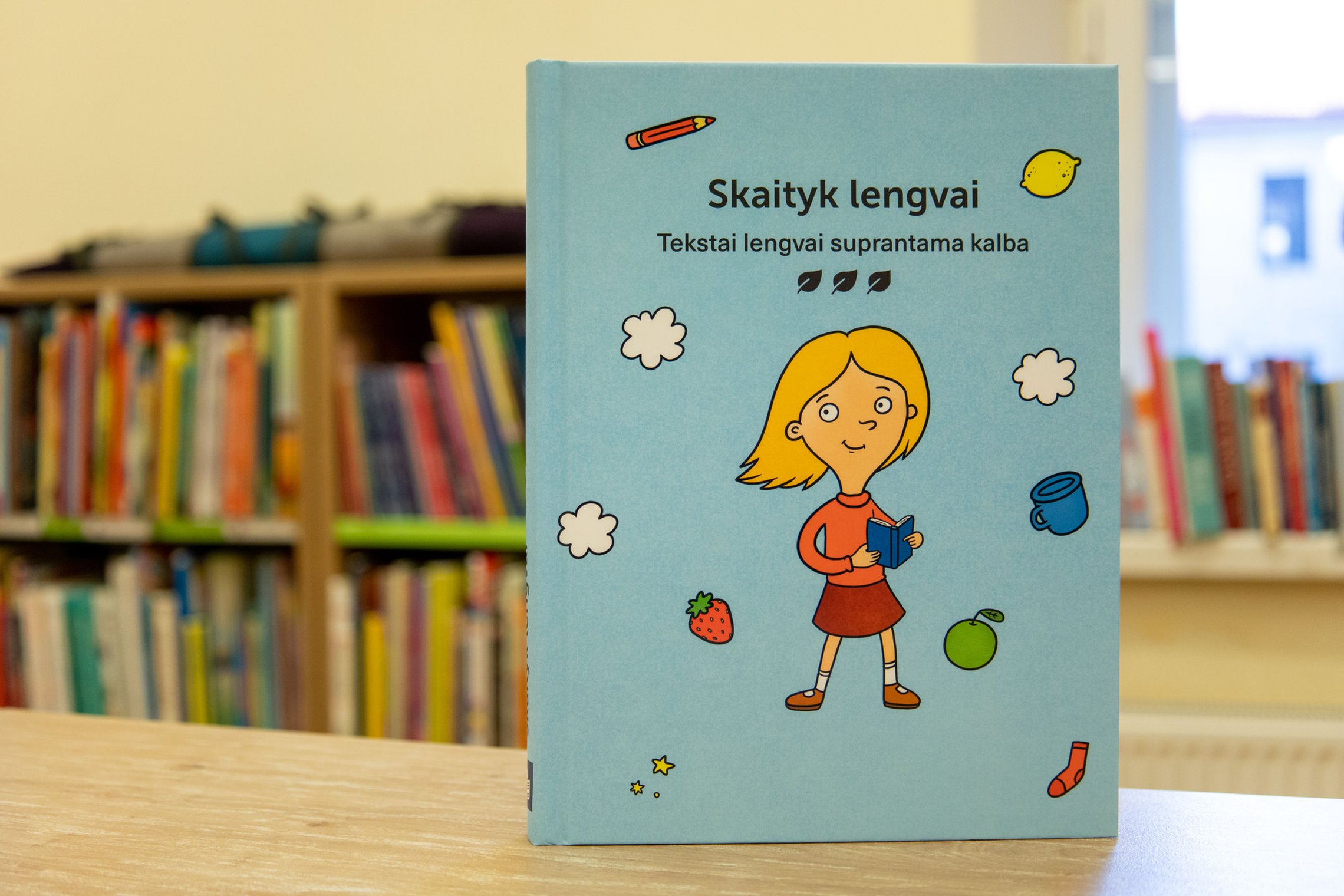 Ant stalo viršeliu į priekį pastatyta knyga Skaityk lengvai, ji papuošta mergaitės, rankose laikančios knygelę, figūrėle, aplink kurią skrajoja įvairūs objektai: debesėliai, obuolys, puodelis, braškė, pieštukas.