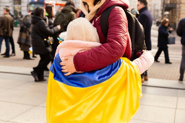 Vaikas apsigaubęs nugarą Ukrainos vėliava stovi lauke apsikabinęs suaugusį žmogų. Jų fone stoviniuoja minia žmonių.