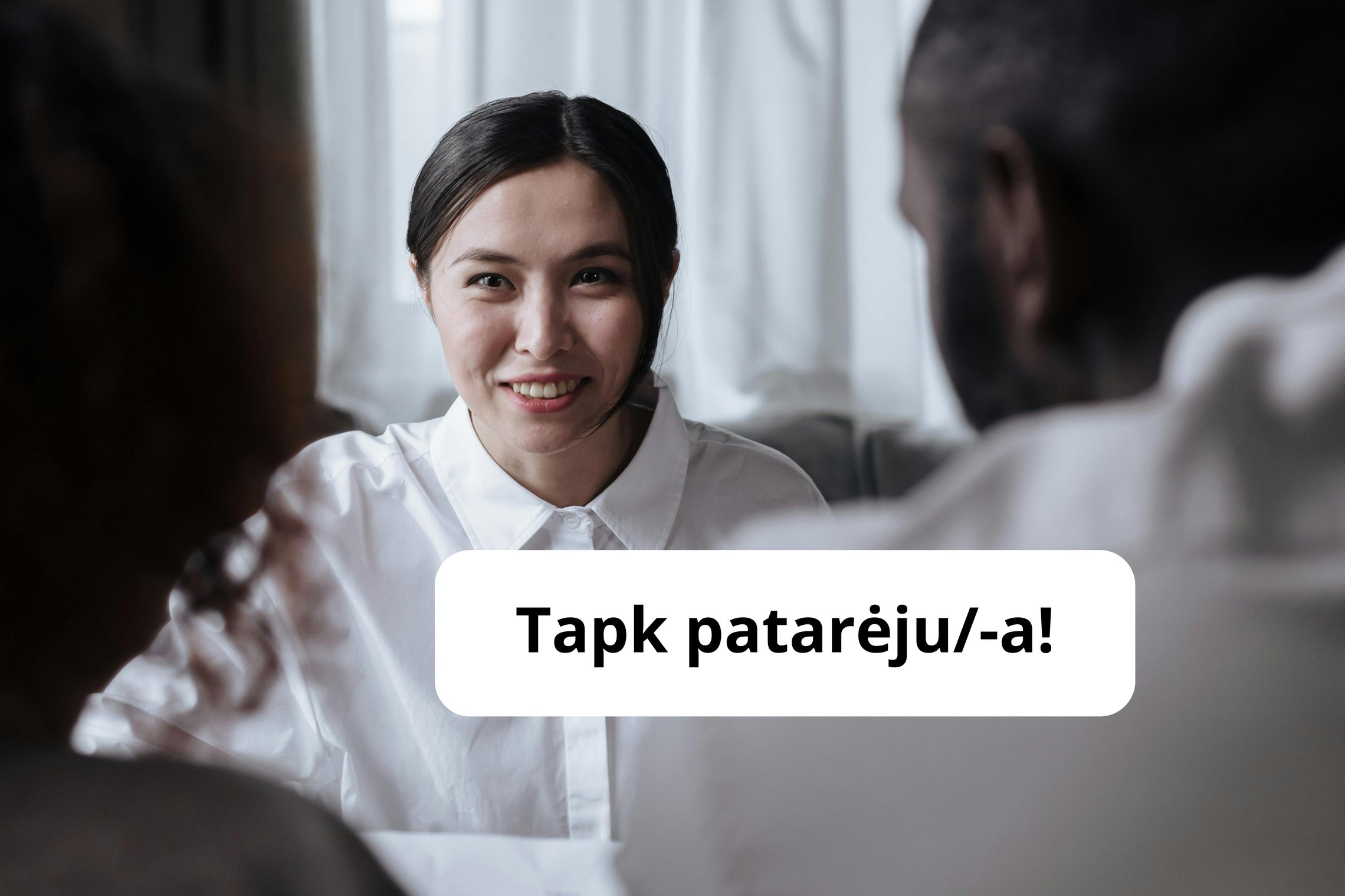 Mergina žiūri į priekį, ji yra su baltais marškiniais, tvarkingai surištais juodais plaukais. Į ją žiūri du žmonės, jie nėra fokuse, matosi tik jų pečiai ir tai, kad jie yra atsisukę į ją. Ant nuotraukos parašyta Tapk patarėju/-a!