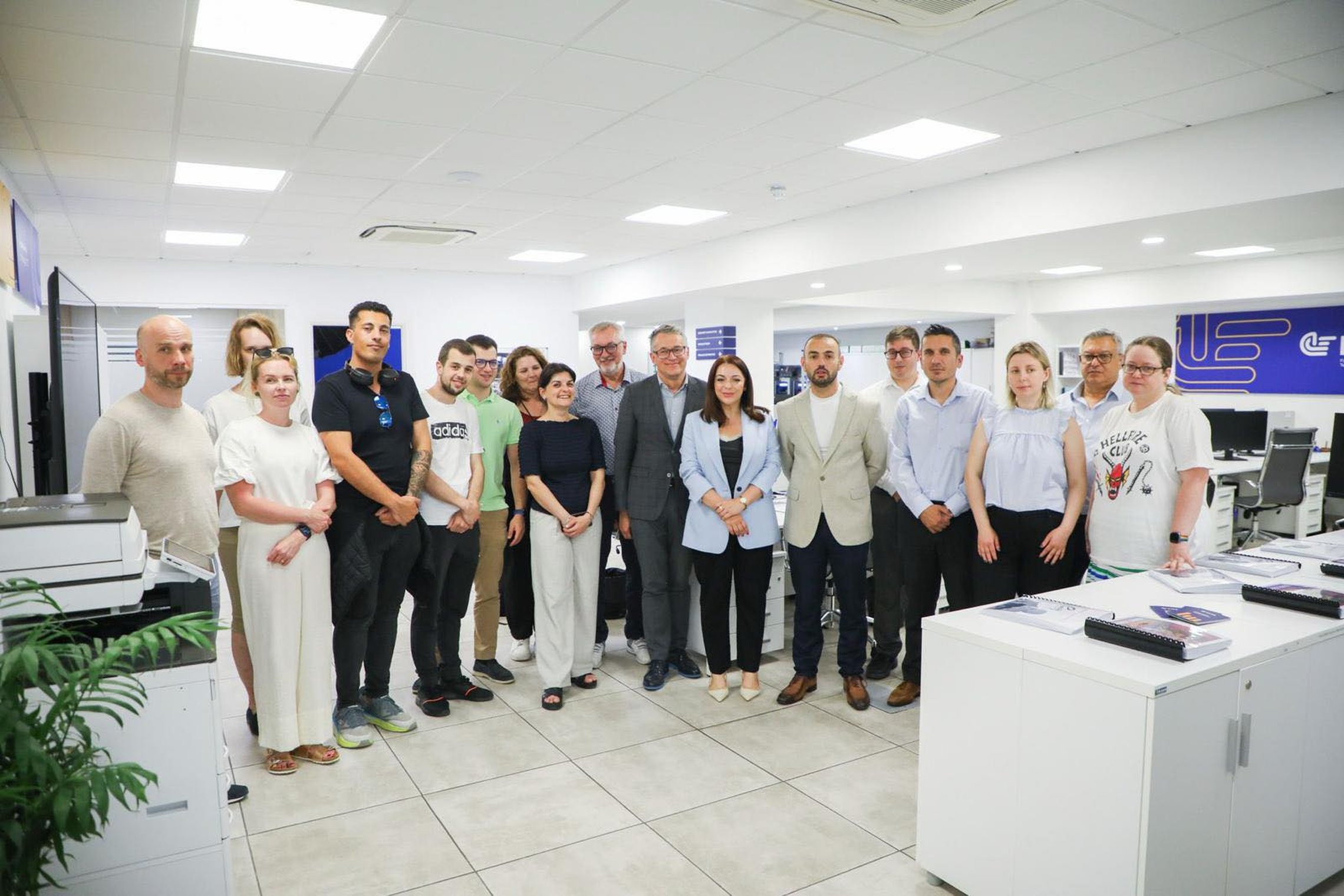 Organizacijos „FITA“ darbo patalpose stovi Erasmus projekto partneriai ir Maltos įtraukties, savanoriškų organizacijų ir vartotojų teisių ministerijos ministrė Julia Farrugia, iš viso nuotraukoje 17 žmonių, jie pozuoja nuotraukai. Šalia matyti spausdintuv