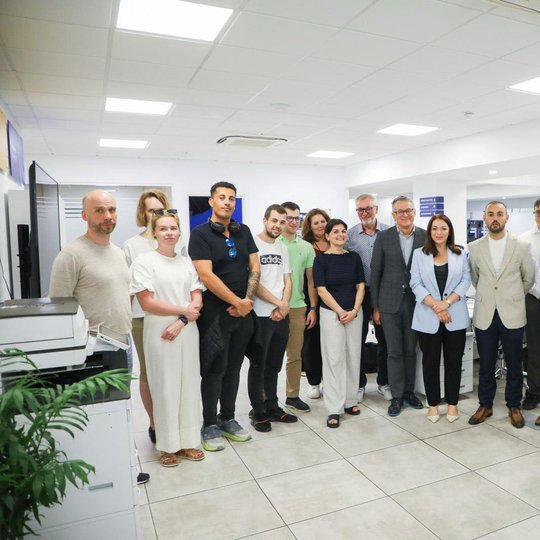 Organizacijos „FITA“ darbo patalpose stovi Erasmus projekto partneriai ir Maltos įtraukties, savanoriškų organizacijų ir vartotojų teisių ministerijos ministrė Julia Farrugia, iš viso nuotraukoje 17 žmonių, jie pozuoja nuotraukai. Šalia matyti spausdintuv