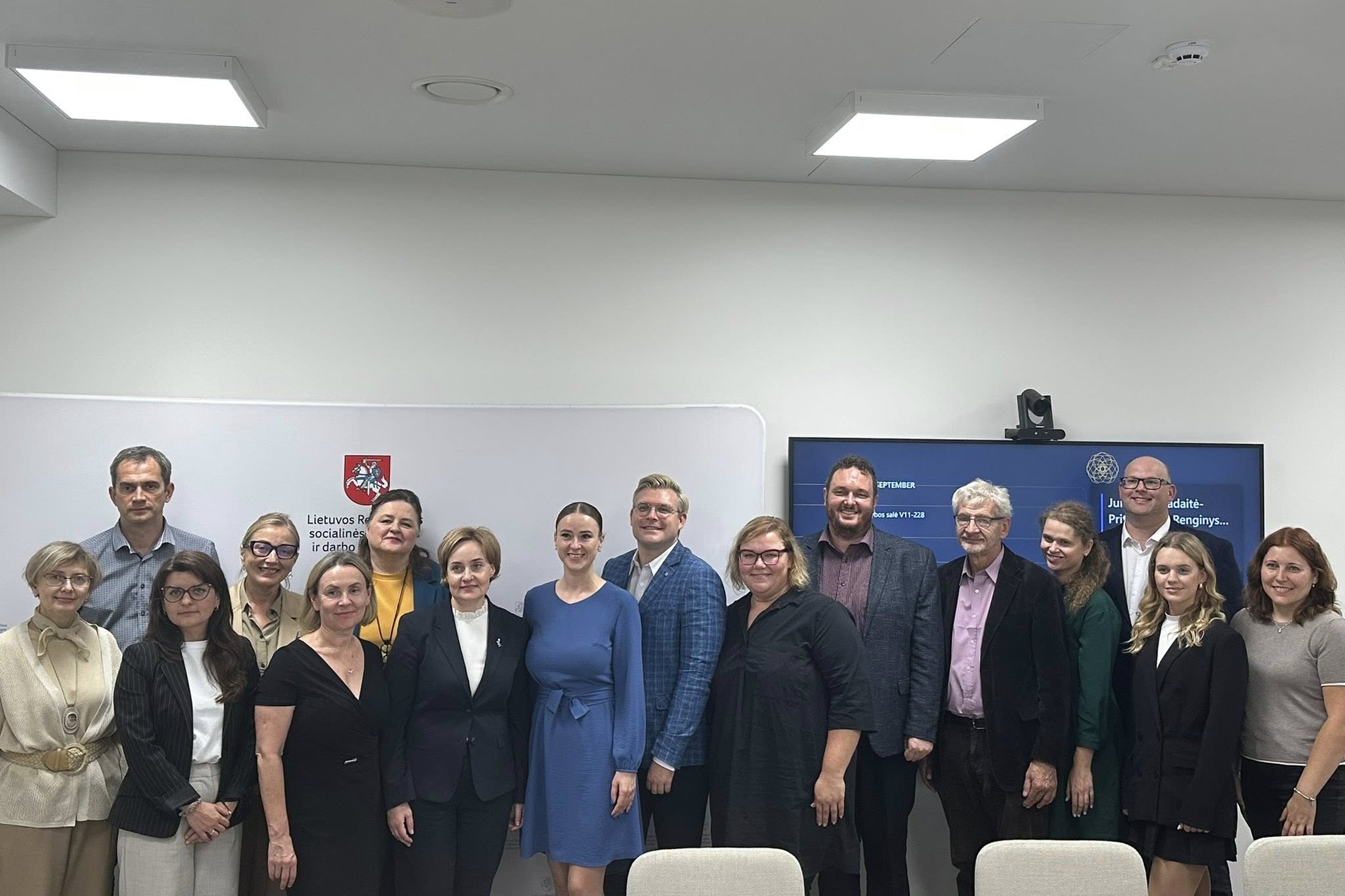 Socialinės apsaugos ir darbo ministerijoje, prie foto sienelės ir skaidrių ekrano, keliomis eilėmis stovi įvairių įstaigų atstovai, dėvi oficialius rūbus, švarkus, marškinius, klasikines sukneles ar kelnes.