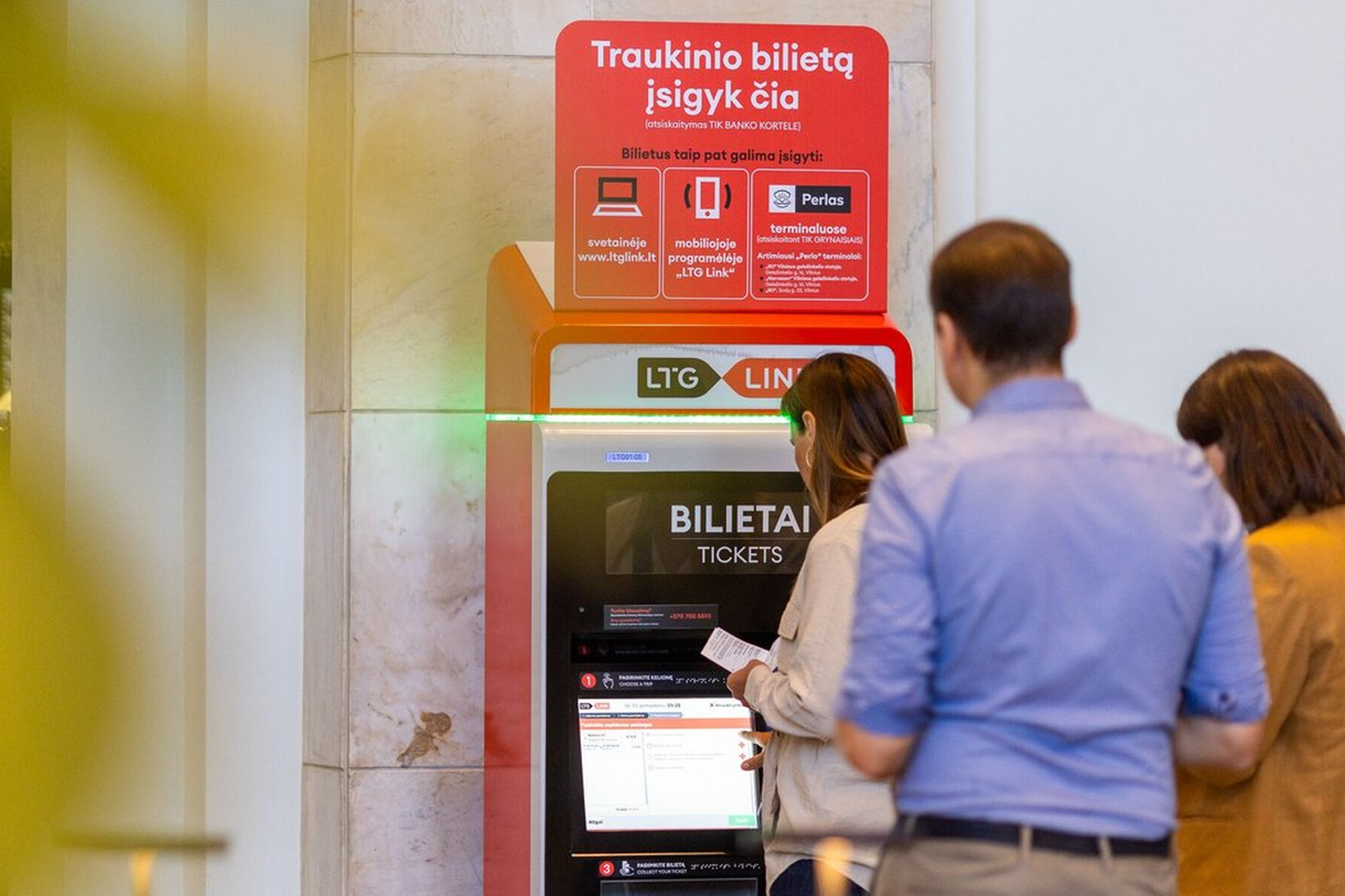 Prie bilietomato traukinių stotyje stovi eilė žmonių, arčiausiai stovi moteris, atsispausdinusi bilietą.