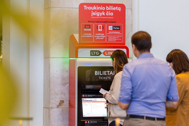 Prie bilietomato traukinių stotyje stovi eilė žmonių, arčiausiai stovi moteris, atsispausdinusi bilietą.