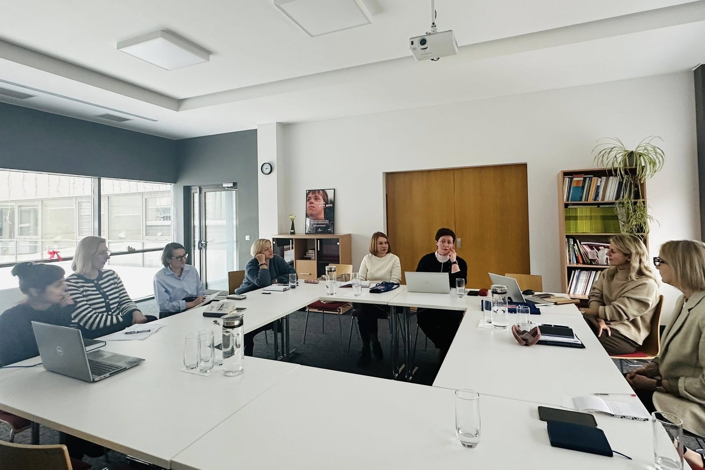 Salėje aplink kvadratinį konferencijų stalą, su ertme per vidurį, sėdi įvairios UŽT ir ANTA specialistės. Ant stalo padėtos stiklinės vandens, užrašinės, kai kurios specialistės turi kompiuterius