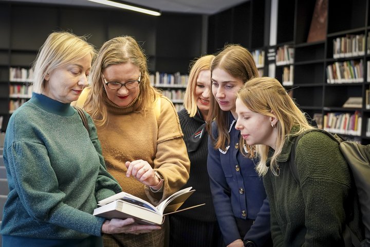 Penkios moterys žvelgia į atverstą knygą, palinkusios link jos, tyrinėja tai, kas ten parašyta. Knygą laiko Agentūros vadovė Eglė, esanti kairėje. Už moterų yra knygų lentynos.