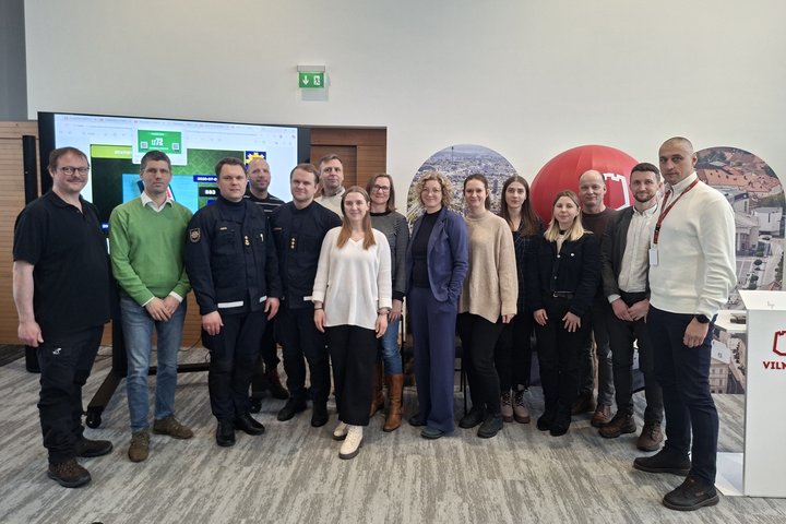Bendrai nuotraukai susibūrę renginio dalyviai, vyrai ir moterys, švelniai šypsosi. Du vyrai atstovauja Valstybinę priešgaisrinę gelbėjimo tarnybą, todėl jie dėvi tamsiai mėlynas uniformas. 