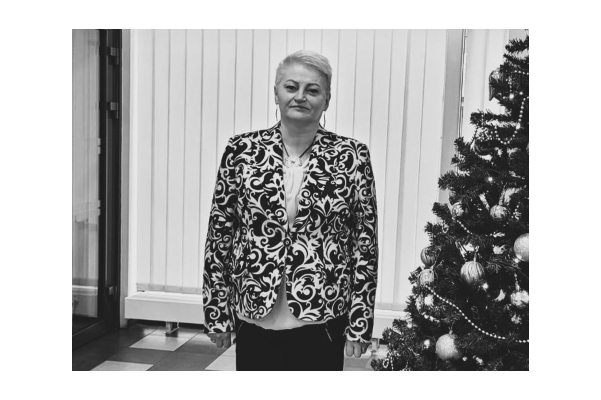 Juodai balta nuotrauka. Moteris trumpais šviesiais plaukais, margu švarkeliu ir juodomis kelnėmis. Švelniai šypsosi. Šalia jos stovi Kalėdinė eglutė.
