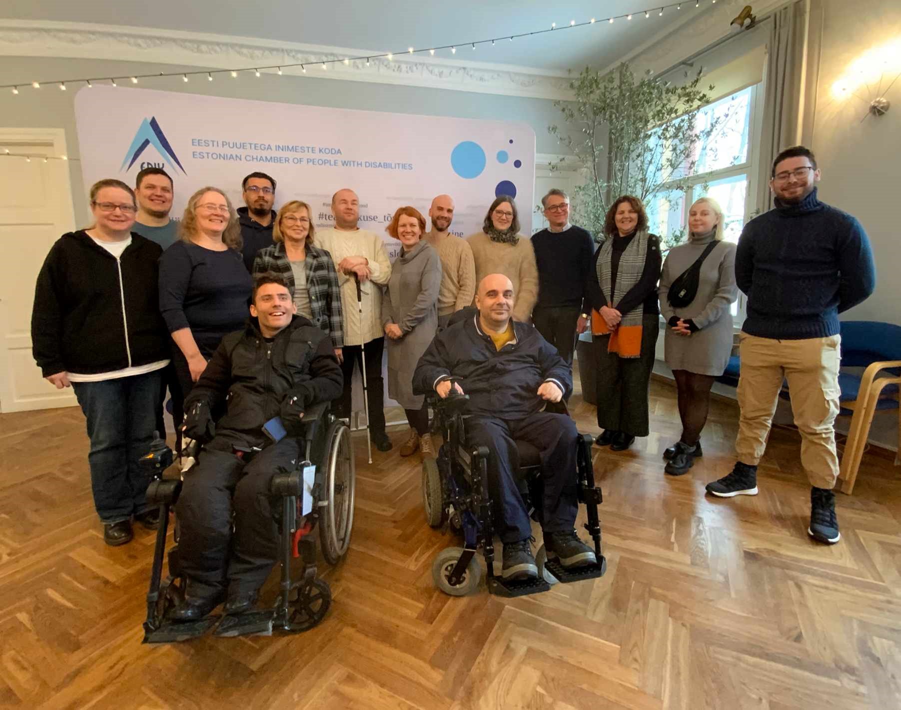 Salėje prie stendo su įvairiais logotipais stovi 15 žmonių, o priekyje yra du žmonės vežimėliuose. Tai Erasmus+ projekto „Digital for all“ partneriai iš Lietuvos, Estijos, Maltos ir Europos autistiškų žmonių asociacijos. 