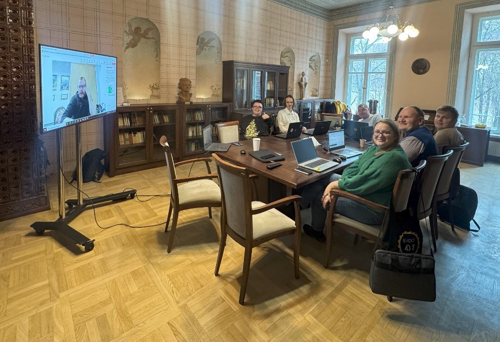 Bibliotekos salėje viduryje yra stalas, prie jo sėdi 6 žmonės –projekto bandomųjų mokymų dalyviai. Ant stalo yra padėti kompiuteriai, telefonai. Netoli stalo yra ekranas, kuriame matosi kitas dalyvis, kuris prie susitikimo prisijungė nuotoliniu būdu. Visi žmonės šypsosi, viena moteris, sėdinti prie stalo, rodo liuksą. 