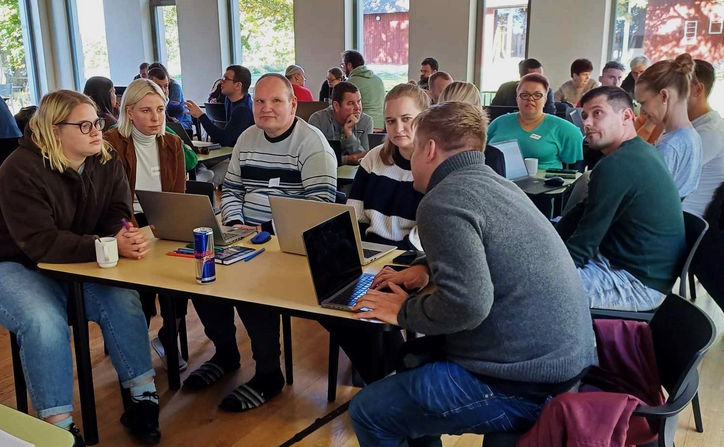 Salėje yra daug stalų, aplink juos sėdi daug žmonių ir kalbasi tarpusavyje. Nuotraukos priekyje prie stalo, ant kurio yra kompiuteriai, telefonai, sėdi Lietuvos bandomųjų mokymų dalyviai ir tarpusavyje kalbasi. 
