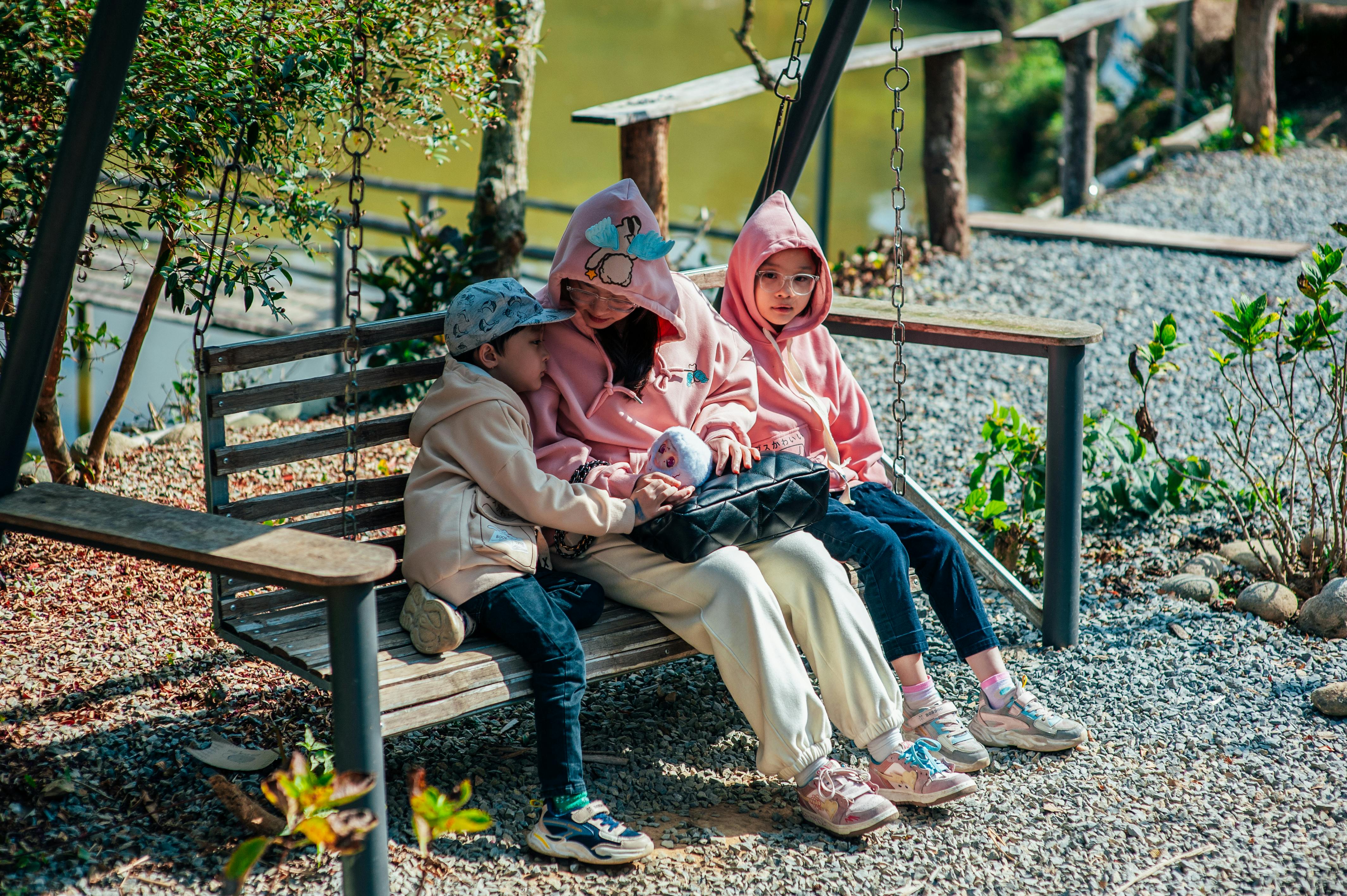 Lauke ant medinio supamo suolo sėdi mama, o iš jos abiejų pusių po vaiką. Kairėje esantis vaikas glaudžiasi prie mamos, yra ją apkabinęs.