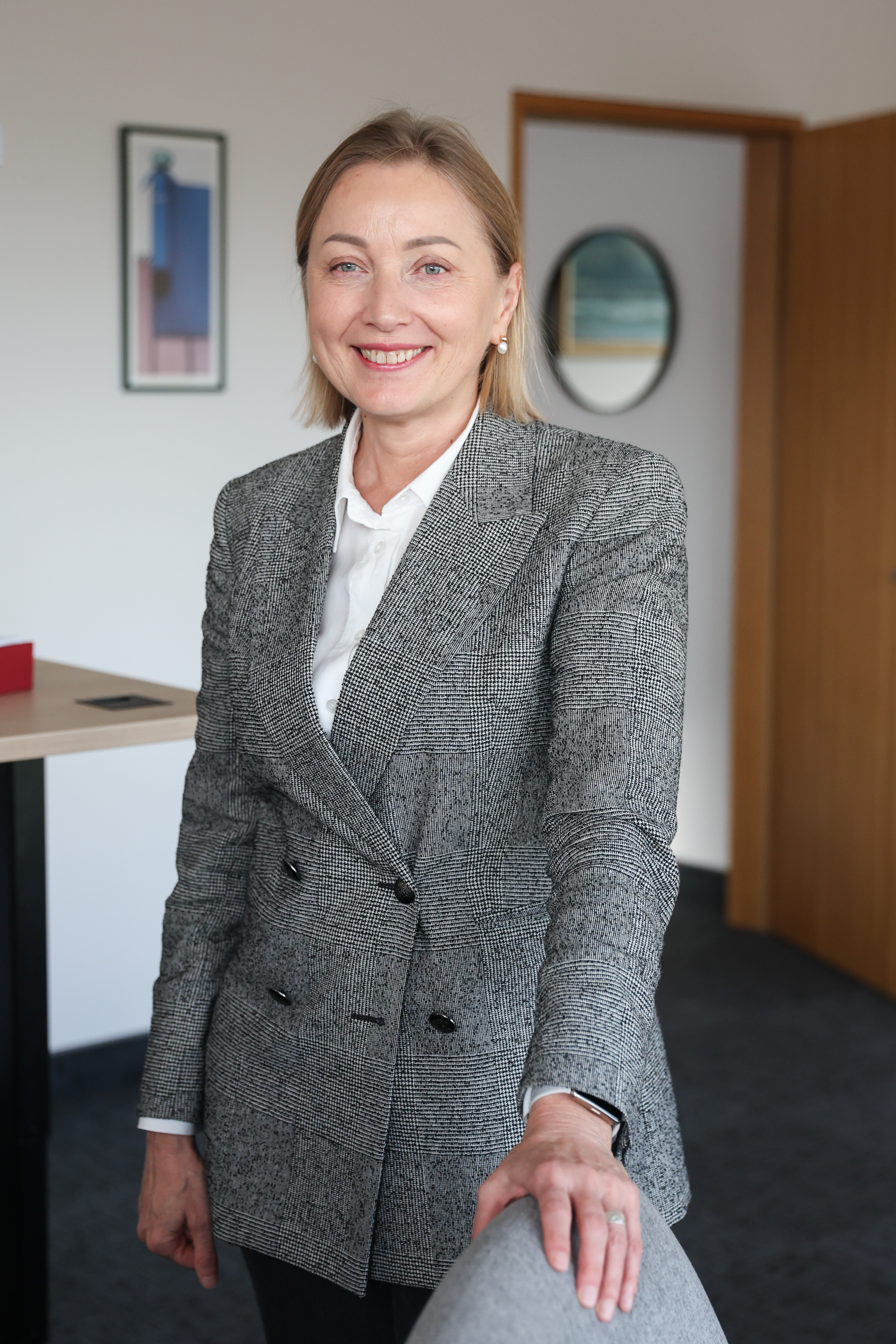 Agentūros vadovė Eglė Čaplikienė stovi kabinete, ji dėvi švarką, šypsosi, viena ranka pasirėmusi į kėdės kraštą.