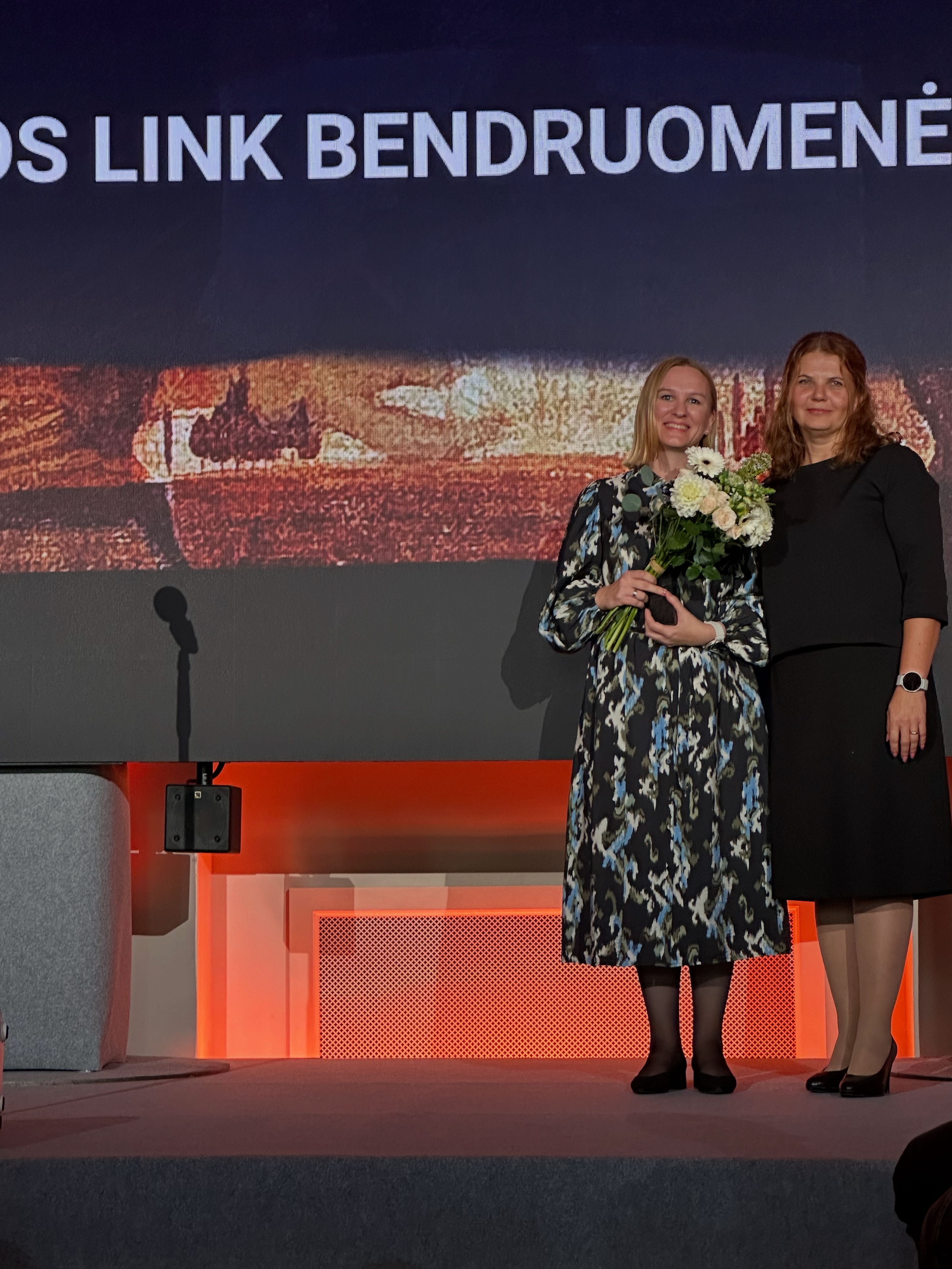 Dvi moterys stovi ant scenos, už jų yra ekranas su Čiurlionio paveikslo motyvais ir dalis užrašo... Link Bendruomenės. Kairėje esanti moteris laiko gėles ir statulėlę, ji pasipuošusi marga suknele. Dešinėje arti prisiglaudusi kita moteris, įteikusi apdovanojimą.