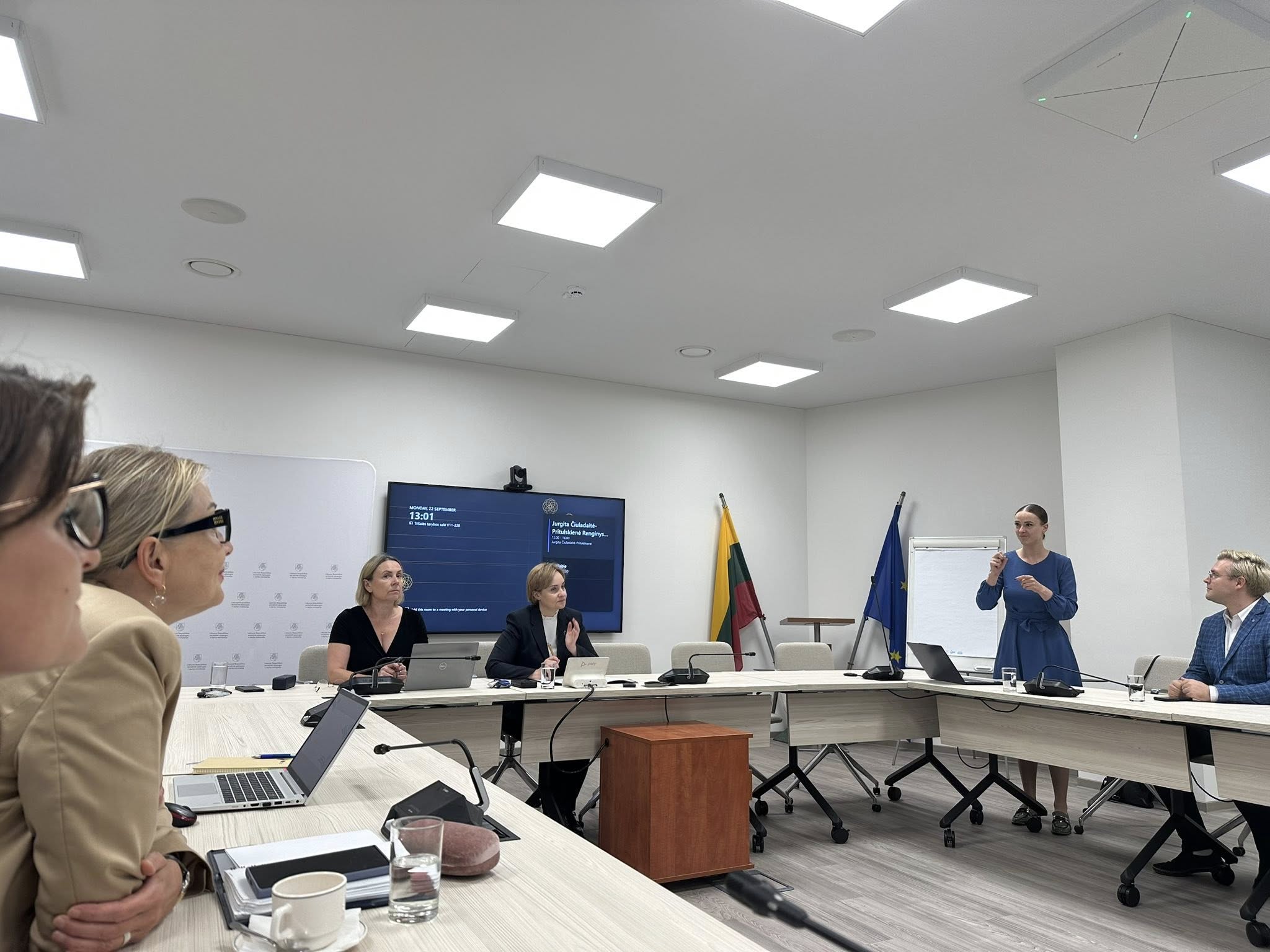 Prie kvadratinio stalo sėdi žmonės, dešinėje stovinti moteris iš vietos kalba gestais, kiti stebi ir skaito iš rankų arba klauso už nugaros esančio vertėjos (nuotraukoje nėra), kuri įgarsina gestais kalbančios Lietuvos kurčiųjų draugijos prezidentės žodžius.