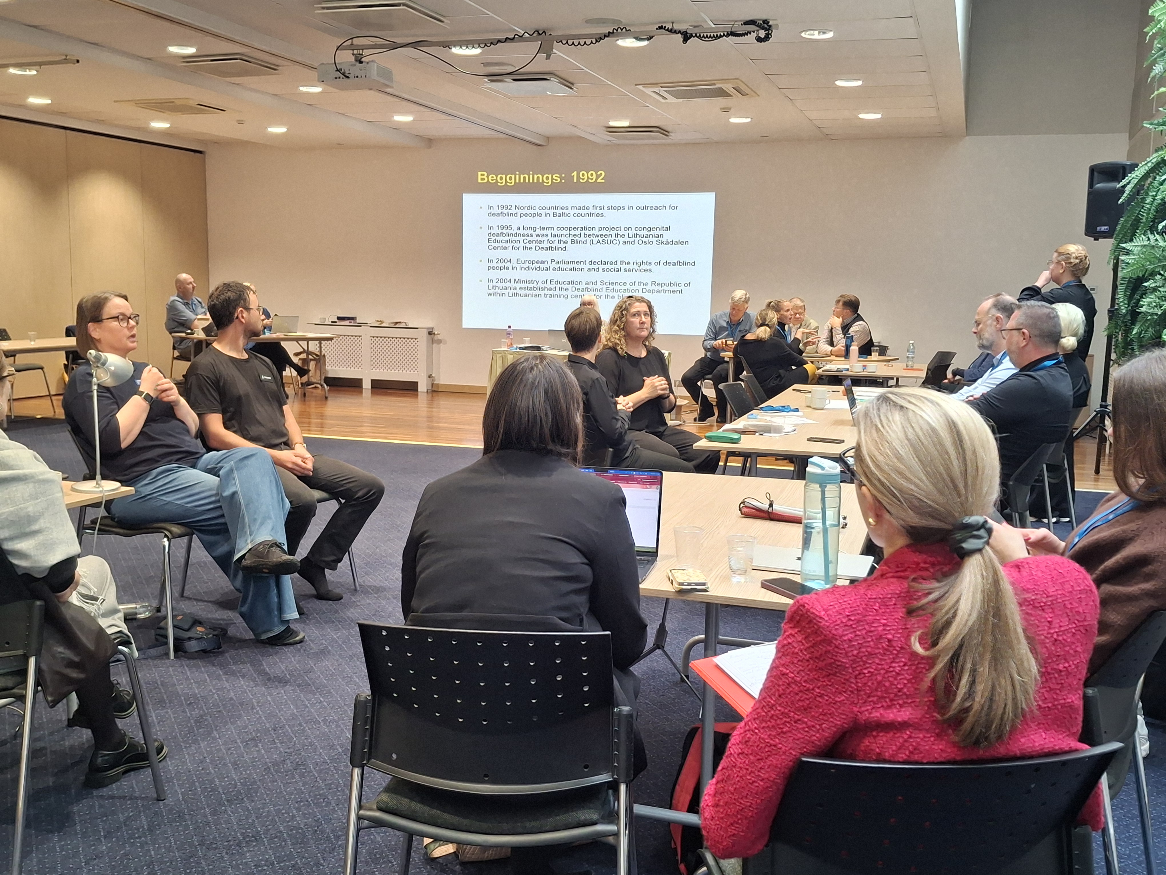 Konferencijų salytėje prie skirtingų pailgų kvadratinių stalų sėdi žmonių grupelės, jos diskutuoja. 