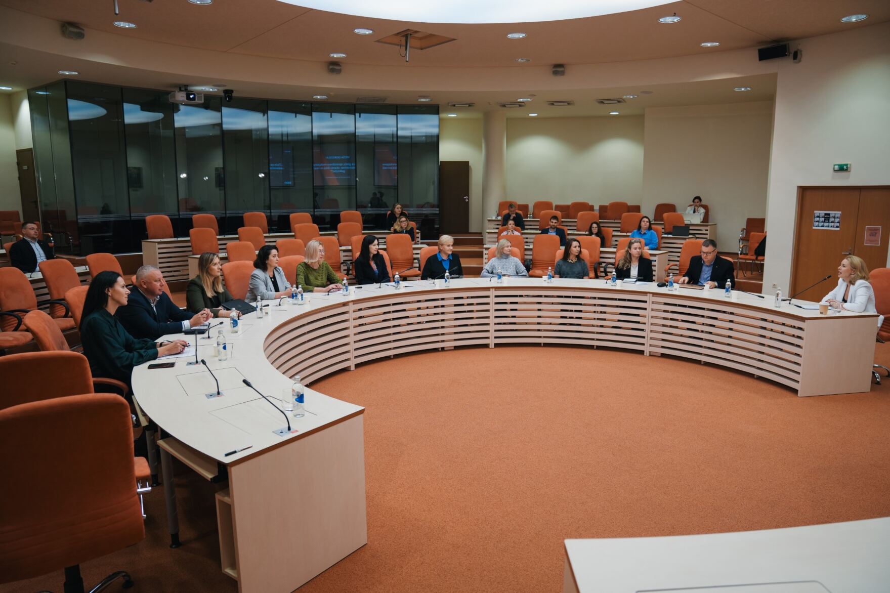 Prie didelio U raidės stalo sėdi įvairių įstaigų atstovai, susirinkę į Mykolo Romerio universiteto auditoriją. Universiteto dvasia atsispindi ir spalvose, kadangi MRU logotipas yra oranžinės spalvos, auditorijos danga ir kėdės yra oranžinės spalvos.