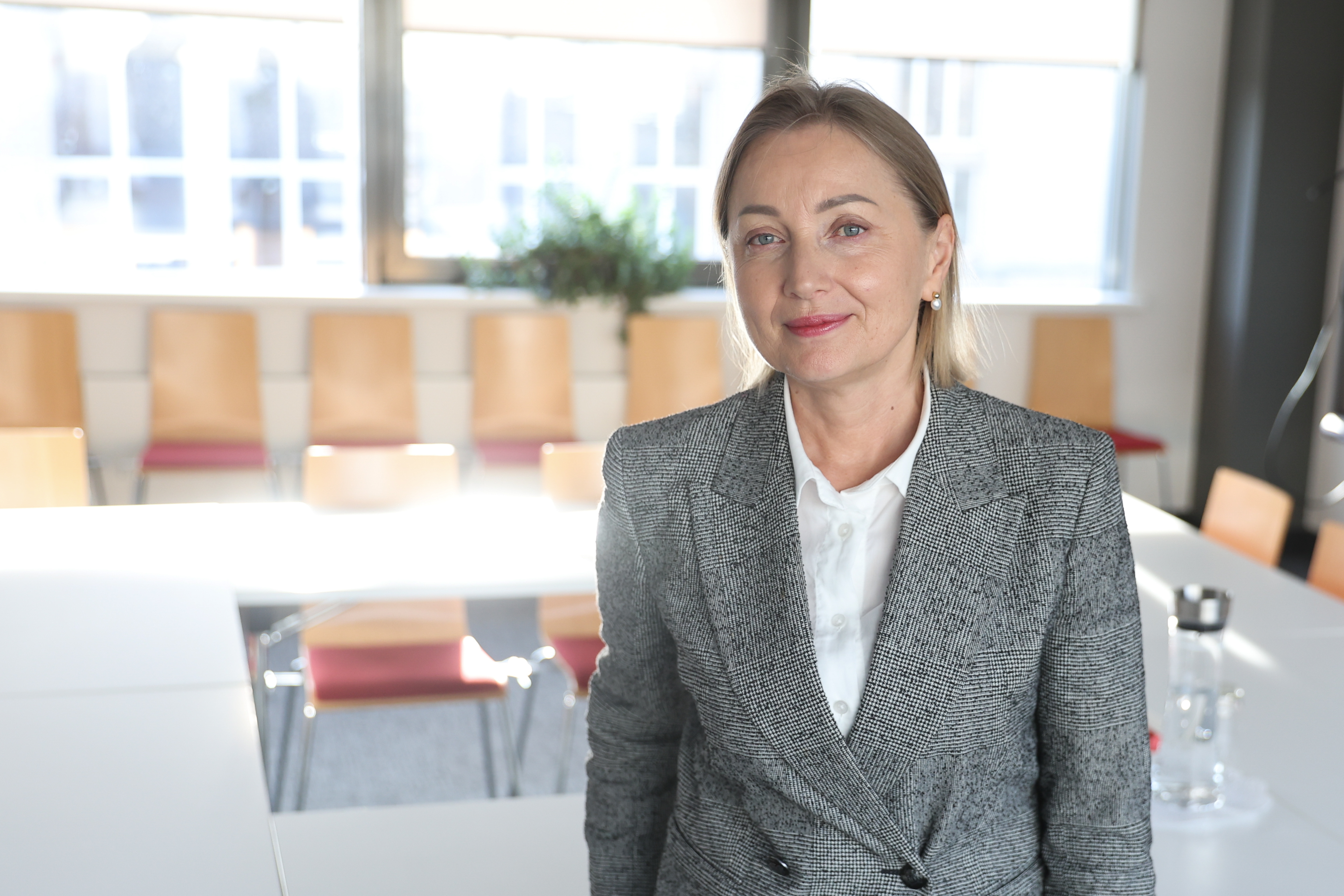 Agentūros vadovė Eglė Čaplikienė, šviesiaplaukė moteris, dėvinti pilką švarką, baltus marškinius, pozuoja darbo kabinete