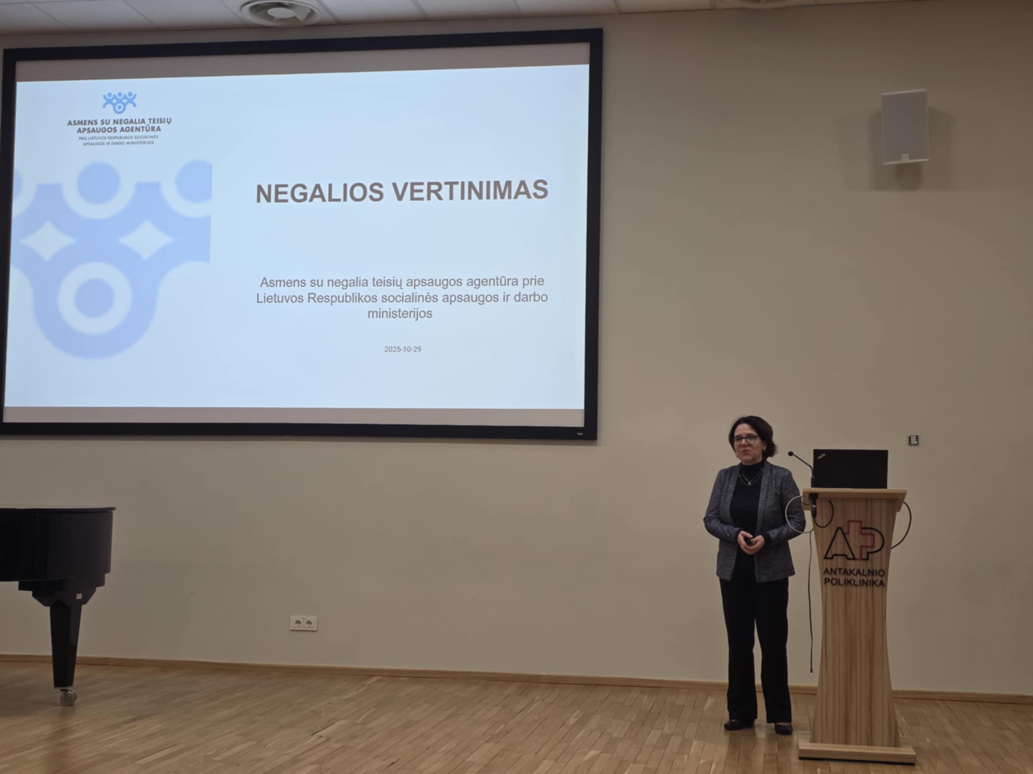 Ant scenos dešinėje stovi pranešėja, esanti greta tribūnos. Jai iš kairės ant didelio ekrano yra skaidrė su užrašu Negalios vertinimas. Žemiau: Asmens su negalia teisių apsaugos agentūra prie Lietuvos respublikos socialinės apsaugos ir darbo ministerijos. Kairėje skaidrės pusėje yra Agentūros logotipas ir grafinis elementas su logotipo dalimi, mėlynais žiedais, kurių viduryje yra mėlyni apskritimai. Ant scenos taip pat matosi dalis fortepijono.