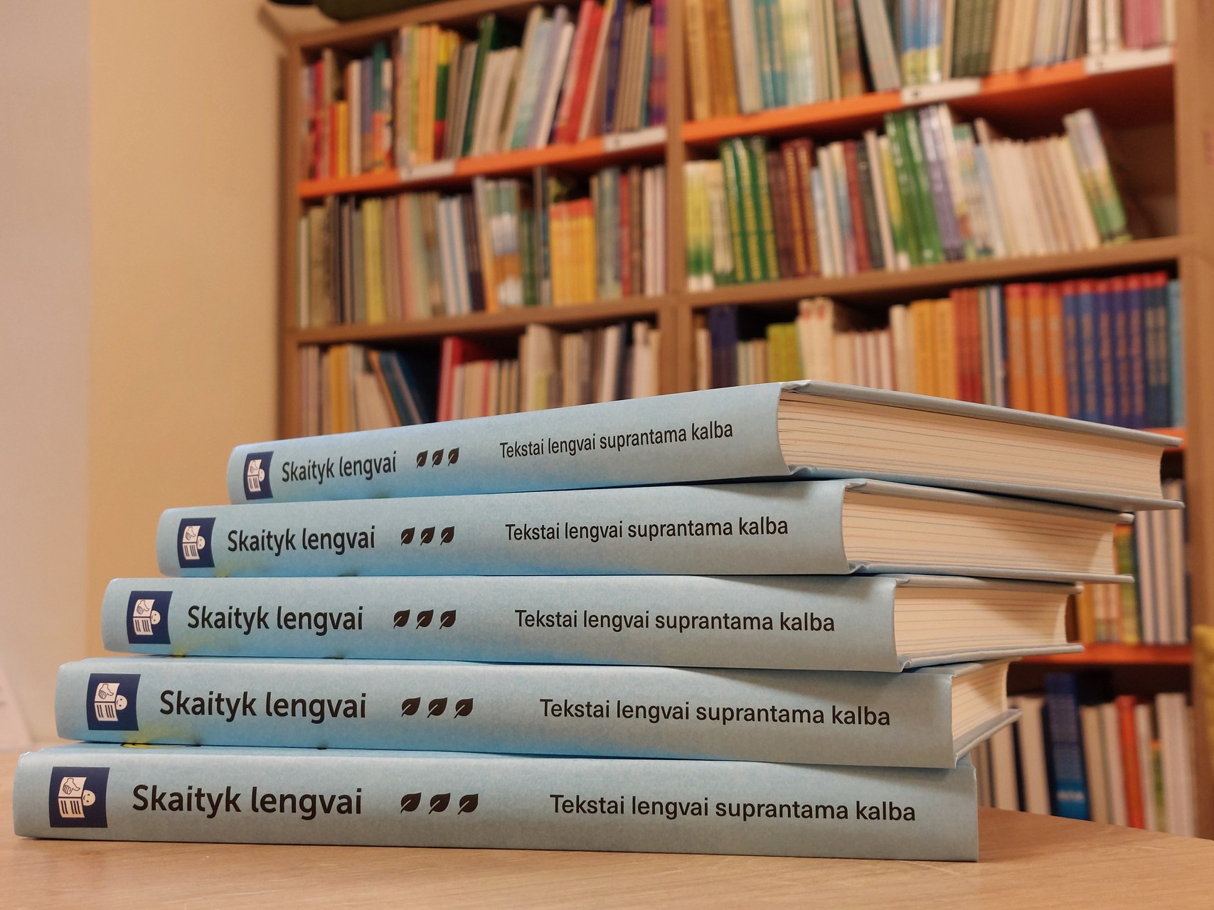 Ant stalo paguldyta krūvelė knygų Skaityk lengvai nugarėlėmis į priekį, už jų tolumoje matosi bibliotekos lentynos, nukrautos įvairiomis knygomis.