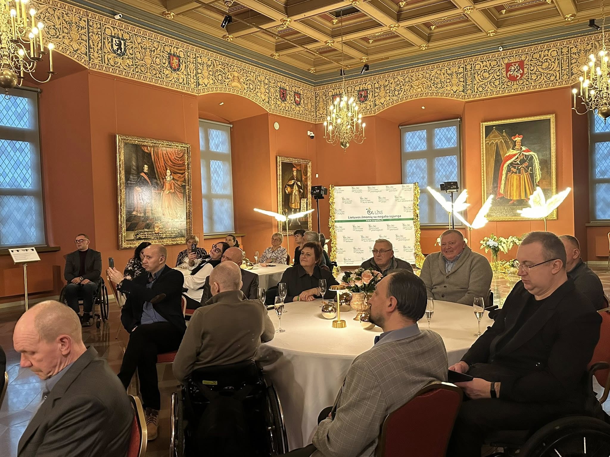 Prie apvalių stalų sėdi organizacijos bičiuliai, partneriai, nariai, klausosi sveikinimo kalbų. Visa erdvė ištaiginga, ant sienų kabo kunigaikščių paveikslai, kabo senoviniai šviestuvai, tai Valdovų rūmų menė.