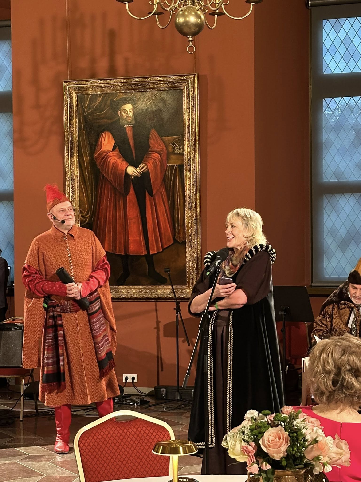 LŽNS pirmoji vadovė Rasa, dešinėje, pasipuošusi ruda valdovės suknia, besišypsanti pasakoja apie organizacijos veiklą ir nueitą kelią. Jai iš kairės stovi vyras, pasipuošęs oranžiniu kostiumu, per juosmenį užsirišęs margą skarą, dėvi pailgą kepurę ir ilgus raudonus batus, jis yra renginio vedėjas.
