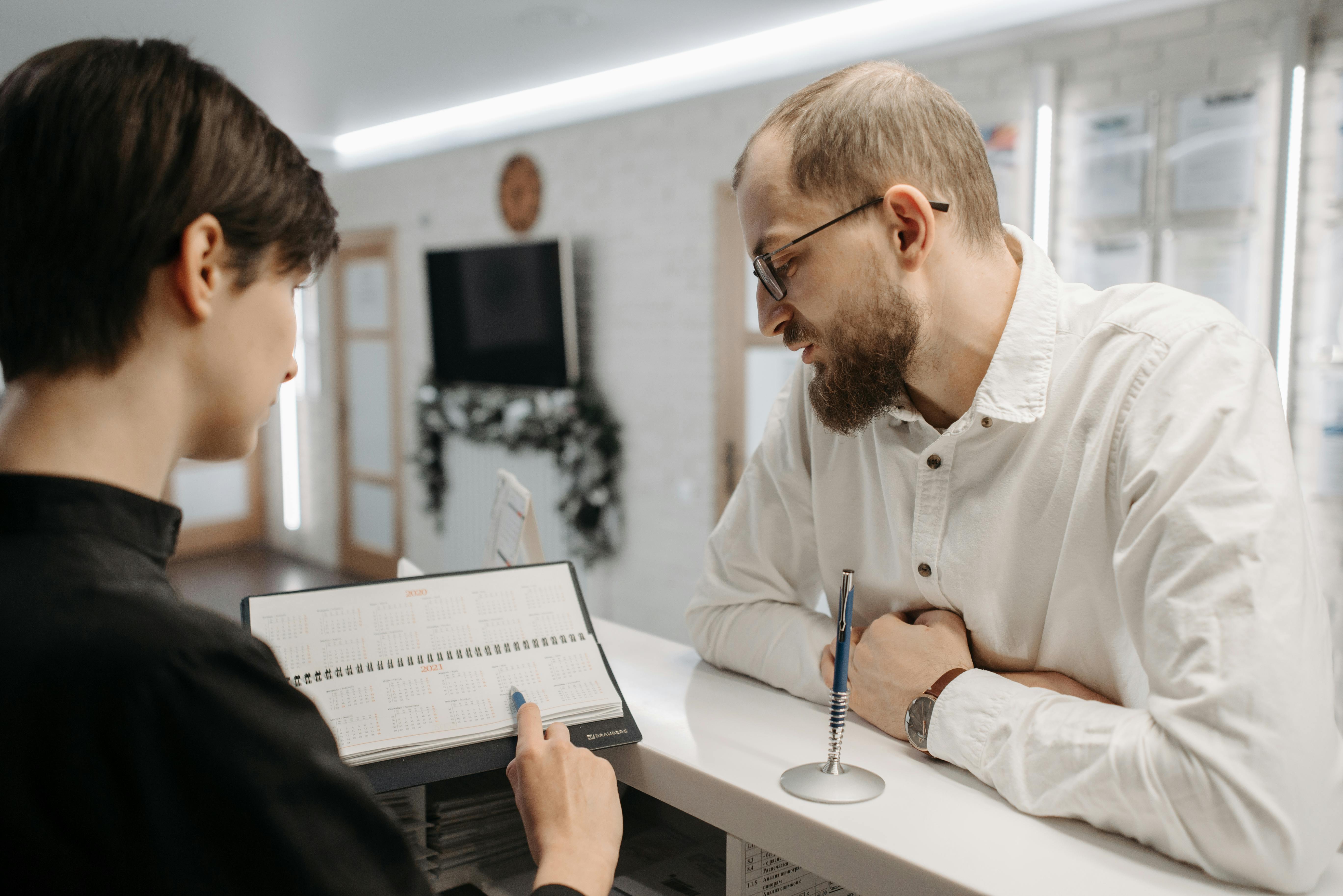 Vidutinio amžiaus vyras prie registratūros stalo, prieš jį stovi specialistė, rankose laikanti kalendorių, tušinuku rodanti į ateityje esančią datą. 