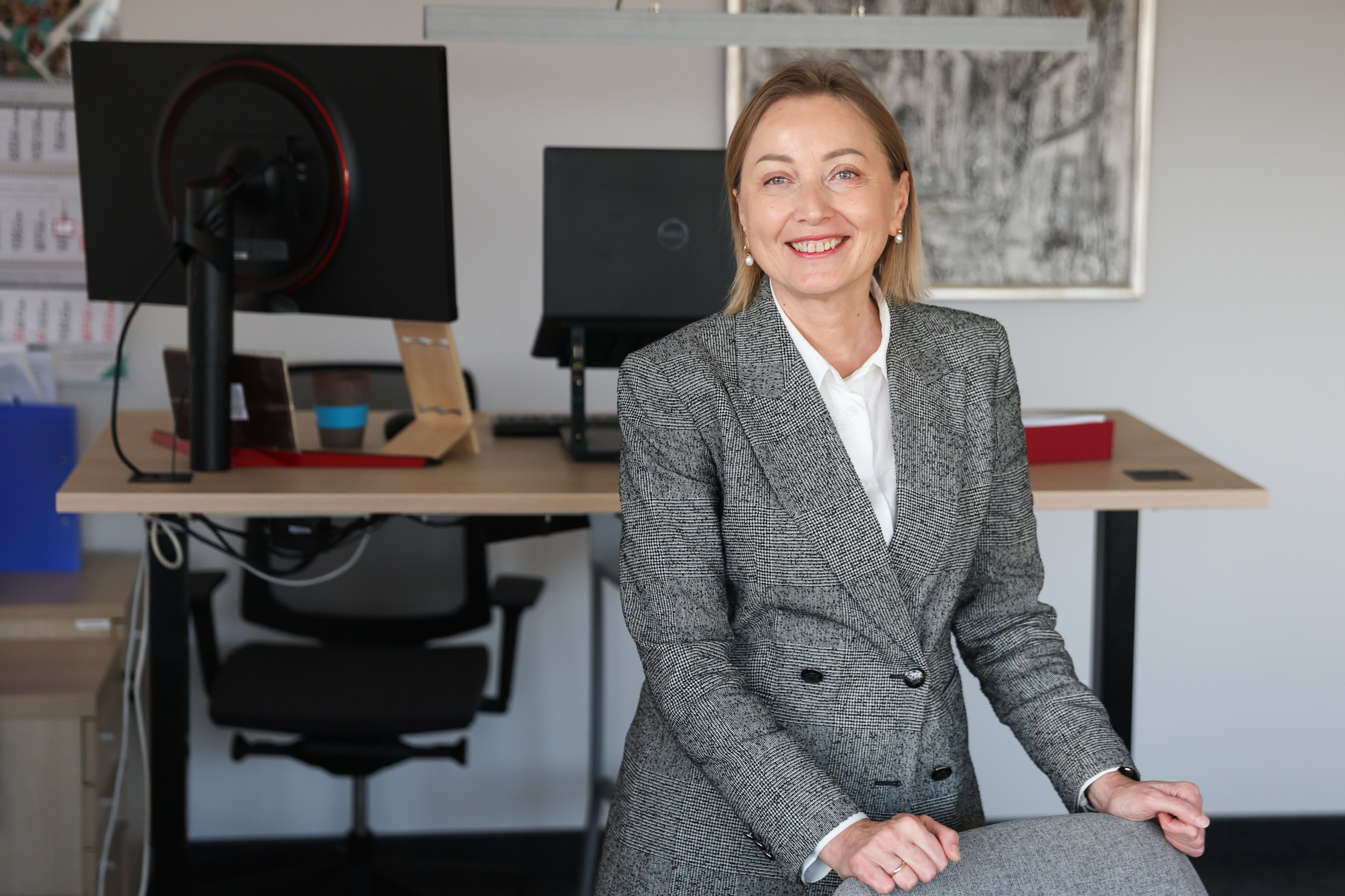 Agentūros vadovė Eglė Čaplikienė savo kabinete, stovi rankomis atsirėmusi į kėdės atlošą, šypsosi, dėvi pilką raštuotą švarkelį ir baltus marškinius. Už jos yra jos darbo stalas, kompiuteris.